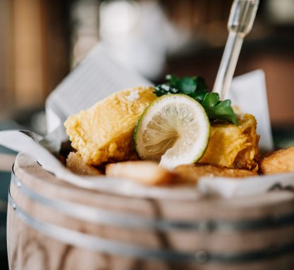 Knusprig gebackener Fisch und Chips serviert in einem kleinen Holzfass getoppt mit einer Limettenscheibe und Petersilie in der Seitenansicht.