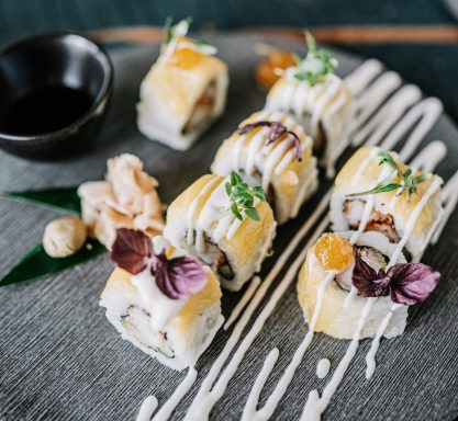 Sieben Sushi-Stücke mit einer Schale Sojasauce auf einer runden, schwarzen Schieferplatte angerichtet und mit Kräutern und Mayonnaise in geschwungenen Linien dekoriert.