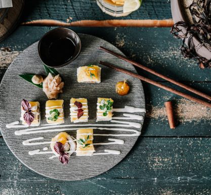 Blick von oben auf sieben Sushi-Stückchen angerichtet auf einer runden Schieferplatte, dekoriert mit Kräutern und Mayonnaise in geschwungenen Linien. Dazu Sojasauce in einem Schälchen und Essstäbchen am Tellerrand.