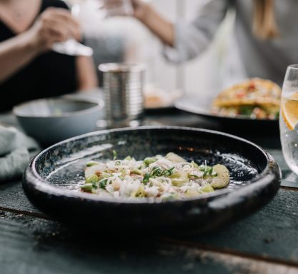 Ceviche aus Kingfish mit Granny Smith und Piment d'espelettes serviert auf einem dunklen Teller mit einem Glas Sprudelwasser mit Orange und Rosmarin.